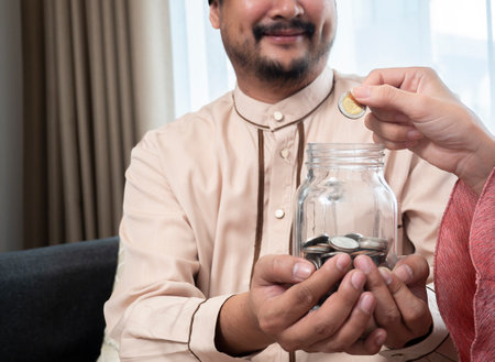 Cropped image of a happy couple at home saving money in a jar glass. Close up hands of a happy Asian muslim couple at home saving money in a jar glass and smilingの写真素材