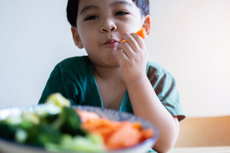 Portrait of cheerful asian boy eating salad with joy. He is looking aside with curiosity and smiling. Kids eating healthy food in kindergarten or at home.の写真素材