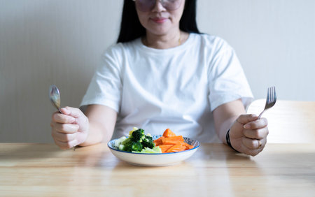 Happy Asian woman eating salad in the simple decorated kitchen and sitting on the table. Healthy food concept.の写真素材
