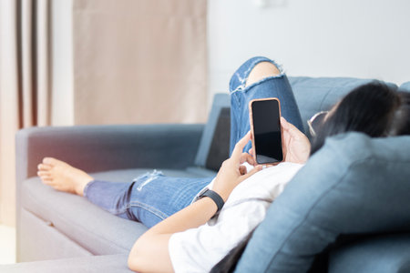 Asian woman enjoys online communication while lying on blue couch in living room. Technology, communication conceptの写真素材