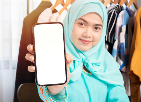 Excited beautiful Asian woman in green sweater and hijab showing blank screen mobile phone isolated over her shop selling through ecommerceの写真素材