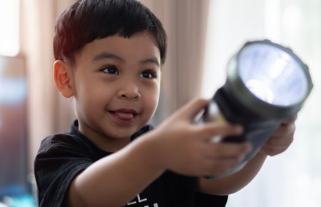Asian toddler in black costume shines a flashlight forward in the room and smiles with joy at what...