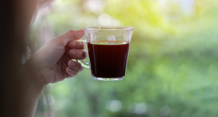 Woman hand holding and showing a glass cup of hot coffee, espresso coffee, black coffee at outdoors on green leaf background. Back view and space for text.の写真素材