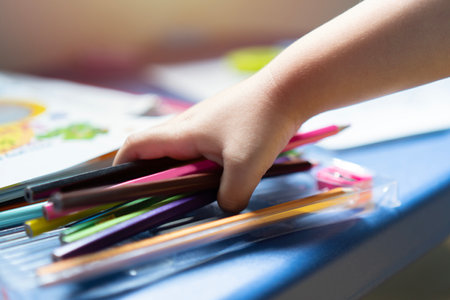 Cropped image of  a little child hand holding a colorful pencils, playing children using pencil crayons  at table indoors. Selective focus. Shot from side.の写真素材