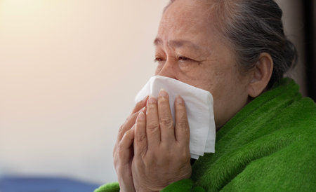 Ill mature woman covered with blanket blowing running nose sneeze in tissue suffer from allergy flu, allergic old lady hold handkerchief got hay fever rhinitis symptom cough at home, allergy conceptの写真素材