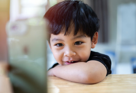 Portrait of cute little Asian resting at wooden table at home. Choosing favorite music or cartoons,  internet, watching video, playing games on smartphone.の写真素材