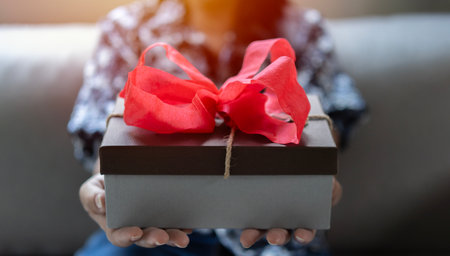 Gift box with red ribbon in the hands of a woman indoor. Shallow depth of field with focus on the box. Gift and give concept.の写真素材