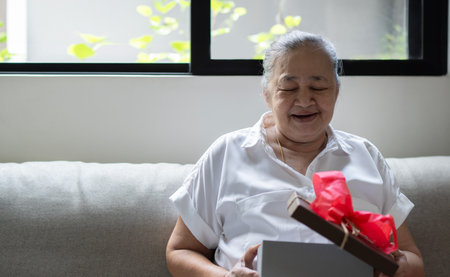 Portrait of a happy smiling Asian older woman opening a gift box. Cropped image of older Asian woman opens box with christmas gift with red ribbon. Selective focus, copy space.の写真素材