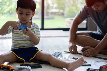 Shot of son sitting nearby the father playing with toy train on house floor. Family weekend entertainment activity at home.の写真素材