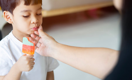 A little Asian boy is eating a ice cream in the summer day at home, and his mom wipes his mouth with a napkin. Sweet eating concept.の写真素材