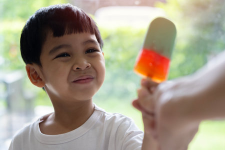 Portrait of smiling little Asian boy with orange ice-cream in hand with blurred background of green...