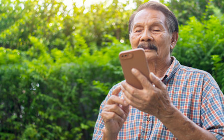 Happy senior Asian man using smartphone and looking at camera while relaxing in the garden. Modern wireless technology easy usage of older generation people conceptの写真素材