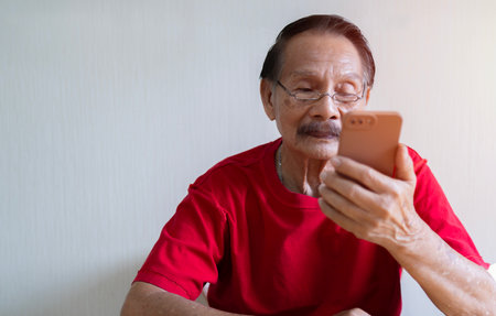 Portrait picture of Asian old man sitting on chair and holding, looking at smart phone screen at home. Retirement age concept.の写真素材