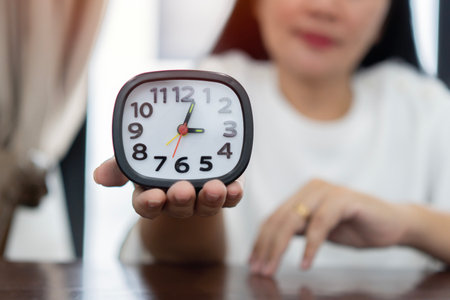 Mature woman hand holding black and white alarm clock at brown wooden table on light room background. Point of view shot. Concept of female biological time.の写真素材
