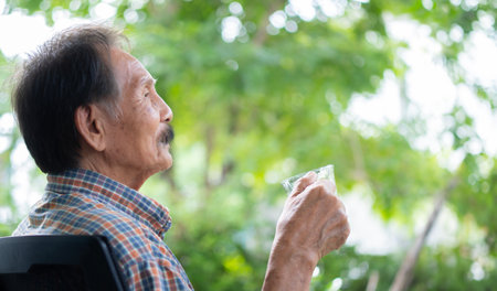 Photo of happy positive elderly Asian man enjoy morning breakfast with a cup of coffee while looking at the green garden at homeの写真素材