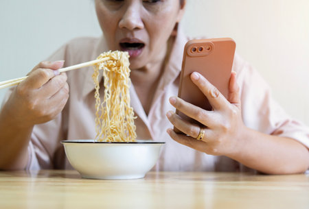 Shot of adult Asian woman eating noodles with chopsticks while watching video on smart phone and sitting on chair at office at home.の写真素材