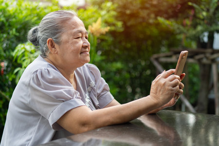Happy old grandmother holding mobile phone, video call with loved ones while relaxing in the garden at home. Technology use in the elderlyの写真素材