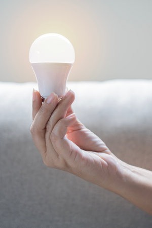Hand holding a LED light bulb on beige sofa background. Using economical and environmentally friendly light bulb concept. Close up, selective focus.の写真素材