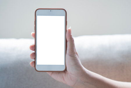 Mockup image of hand holding white mobile phone with blank white screen on light beige sofa background. Phone with blank screen, copy space.の写真素材