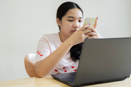 Cropped photo of pretty Asian woman using her smartphone while working on laptop computer and sitting at home, selective focus on handの写真素材