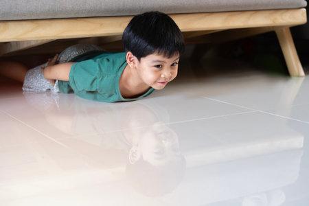 Happy Asian little child boy plays peekaboo or hide and seek under the sofa in the livingroom. Family playing games inside their home.の写真素材