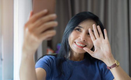 Happy Asian woman waving hand and using smartphone app enjoying online virtual chat video call with friends.の写真素材