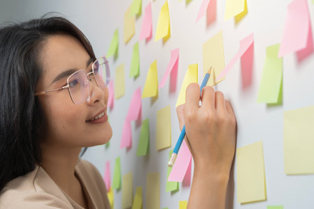 Happy Asian business woman write short note to do list in sticker on wall in meeting office room....