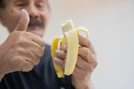 Defocused happy senior man wwith thumb up and isolated on a white background holds a peeled banana in his hand ready to be eatenの写真素材