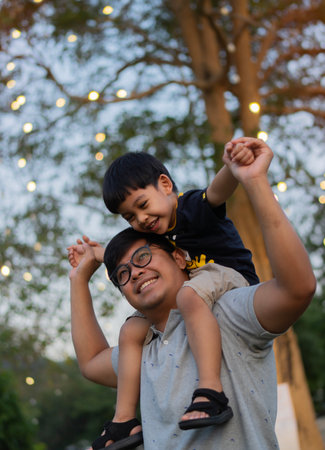Portrait of happy Asian father giving son piggyback ride on his shoulders and looking up over night nature background. Cute boy with dad playing outdoor.の写真素材