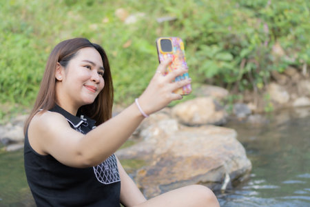 Smiling Asian female taking selfie by smartphone along sunny summer stream. Lifestyle concept.の写真素材