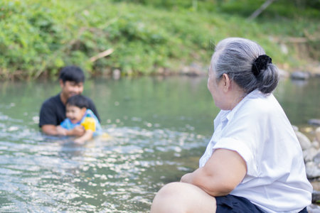Shot view of grandma sits and looks at her son and grandson playing in the river on the summer camping day.の写真素材