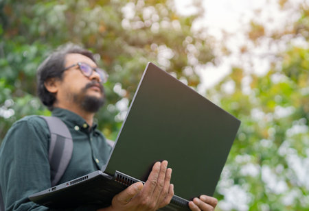 Happy hipster young Asian man wearing glasses standing and working on laptop in the park. Selective focus, copy space.の写真素材