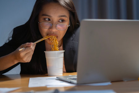 Asian business woman eating instant cup noodle while working on laptop in office at late night. Selective focu.の写真素材