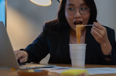 Cropped image of happy Asian businesswoman eating instant noodles when working on report late at night. Selective focus.の写真素材