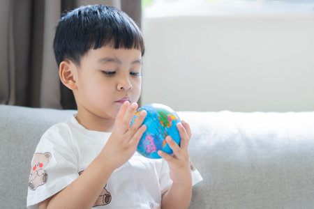 Conceptual photography of a child holding a globe in his hands. Asian little boy sitting on sofa and play globe ball with real sunlight from window at home.の写真素材