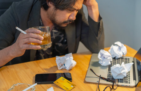Bearded male hold head with hands look down sitting at office feels depressed desperate, drinking strong alcoholic drink, having serious dependence alcoholism. Selective focus.の写真素材