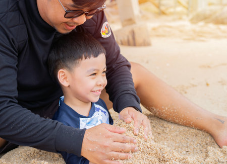 Cropped view of little boy and dad sitting on sandy beach near sea and mountain ridge while having fun during sunny dayの写真素材