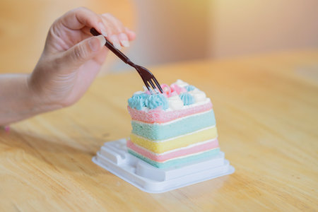 Close-up image of a woman hand eating a painbow cake in plastic tray over wooden table at cafe....