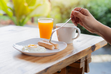 The woman eat by fork with breakfast on the table. American style breakfast with fried eggs, sausage, coffe and juice. The concept of a healthy morning.の写真素材