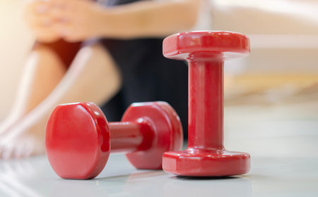 Two red dumbbells on the floor with blurred image of tired exhausted woman in sportswear resting behind. Selective focus.の写真素材