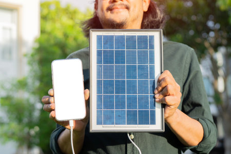 Close-up happy Asian adult man charging mobile phone with portable solar panel on green park background. Isolate screen, copy space.の写真素材