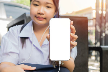Teenage girl in school uniform holding a phone with isolate screen and looking at the camera with earphones outdoors. Copy space.の写真素材