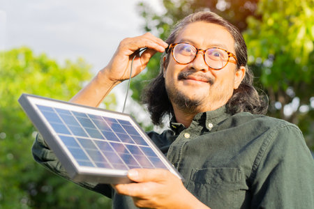 Surprised adult Asian man with portable solar panel charging his own energy on green natural...
