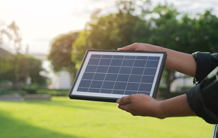 Side view of man holding portable solar panel with reflected sunlight at city park. Alternative energy, renewable energy concept.の写真素材