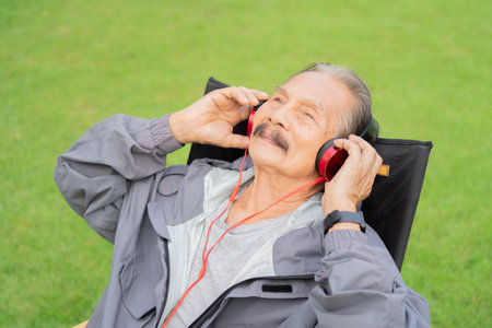 An happy older man is sitting on a camping chair over grassy field, wearing headphones and listen to...