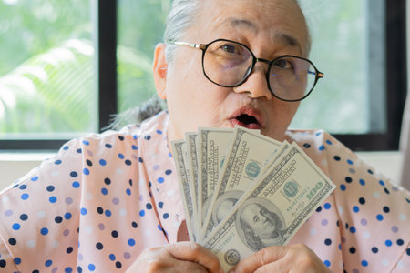 Senior grey-haired Asian woman holding 100 US Dollar banknotes with smiling friendly gesturing...