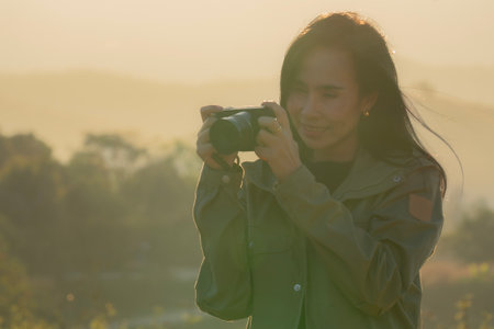 Happy young tourist Asian woman holding a mirrorless camera on the mountain in the evening. Tourist journey trip concept.の写真素材