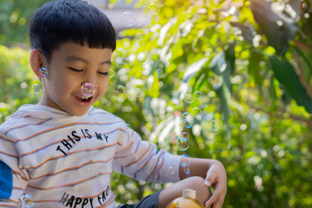 Little Asian child boy among lots of flying bubbles, having fun while playing with rainbow bubbles in the park.の写真素材