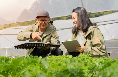 Farmer couple holding laptop and seedling tray to examining quality of vegetables in a greenhouse. Technology Smart Farming and Innovation Agricultural Concepts.の写真素材