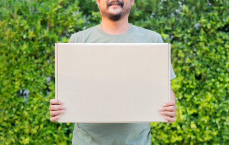 Cropped view of man carrying cardboard box over nature background with copy space. Parcel shipping service or delivery packaging conceptの写真素材
